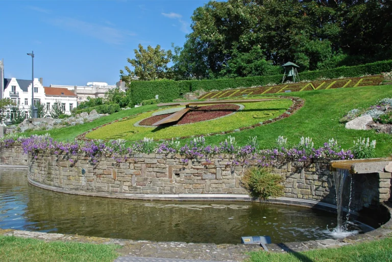The flower clock planted in a sloping lawn above a curved stone pool with flowering borders