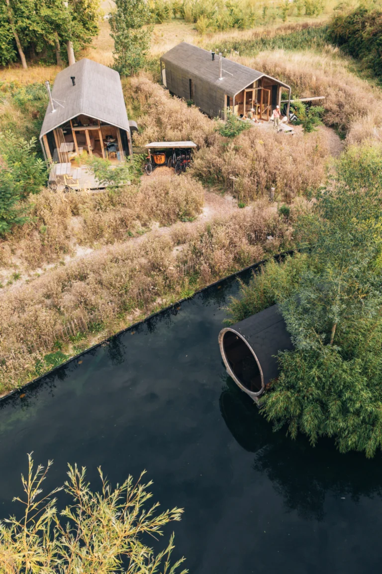 Aerial view of two timber cabins in a wild meadow beside a dark pond with a barrel sauna