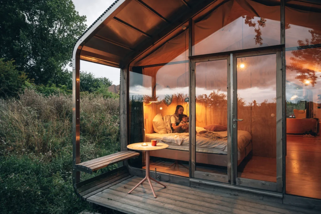 Glass-fronted wooden cabin glowing at dusk, with a small deck and bench among wild grasses
