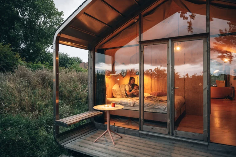 Glass-fronted wooden cabin glowing at dusk, with a small deck and bench among wild grasses