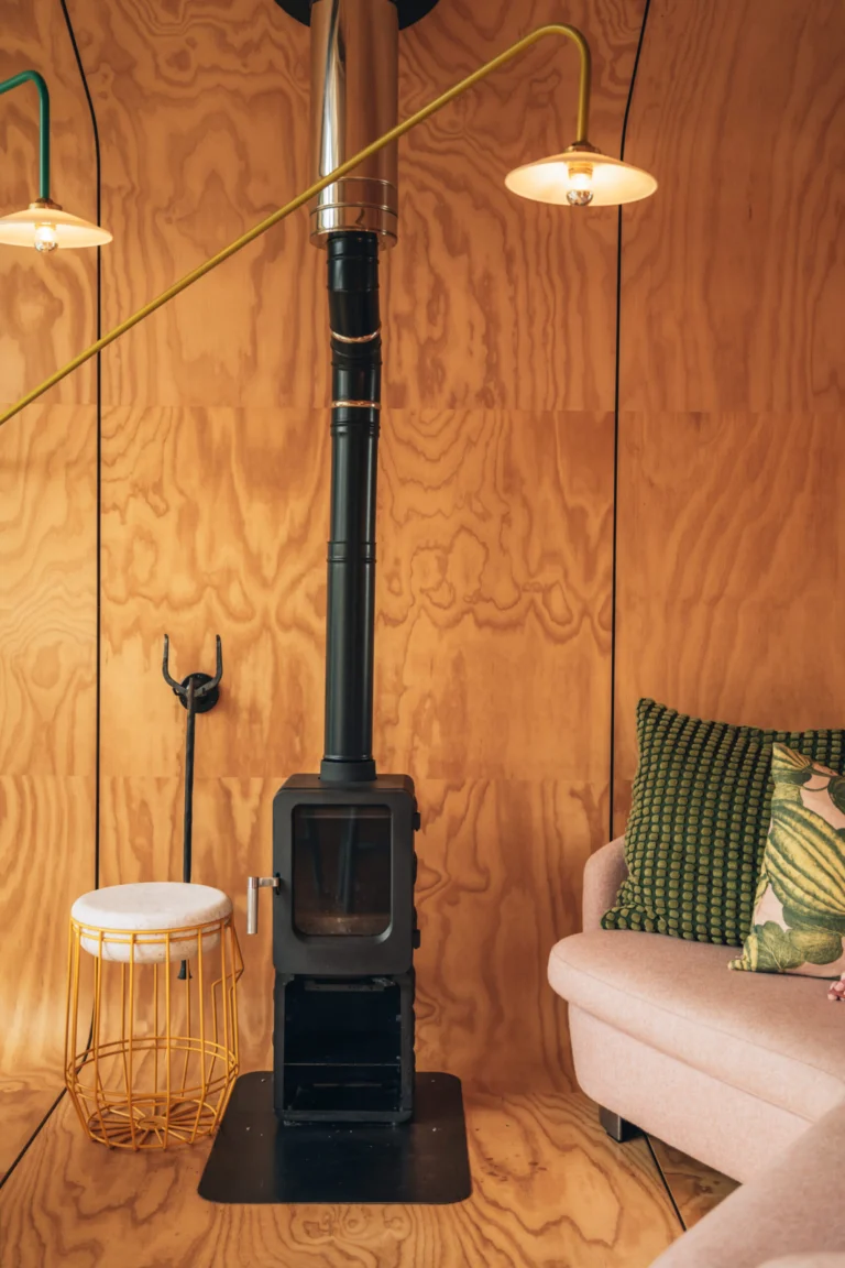 Plywood-lined cabin interior with a black wood-burning stove, pink sofa and brass wall lamps