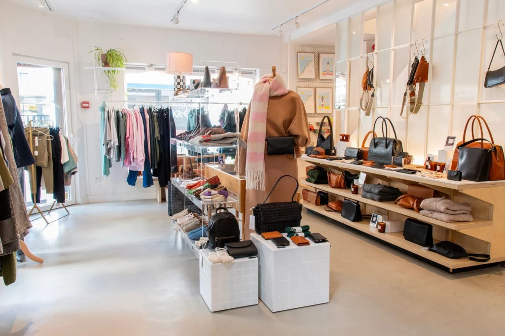 Boutique interior with rails of clothing, a mannequin in a camel coat and pink scarf, and wooden shelves of leather handbags