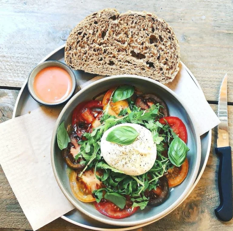 A salad of tomatoes, rocket and basil topped with a ball of soft white cheese, served with a slice of dark bread and a small pot of dip