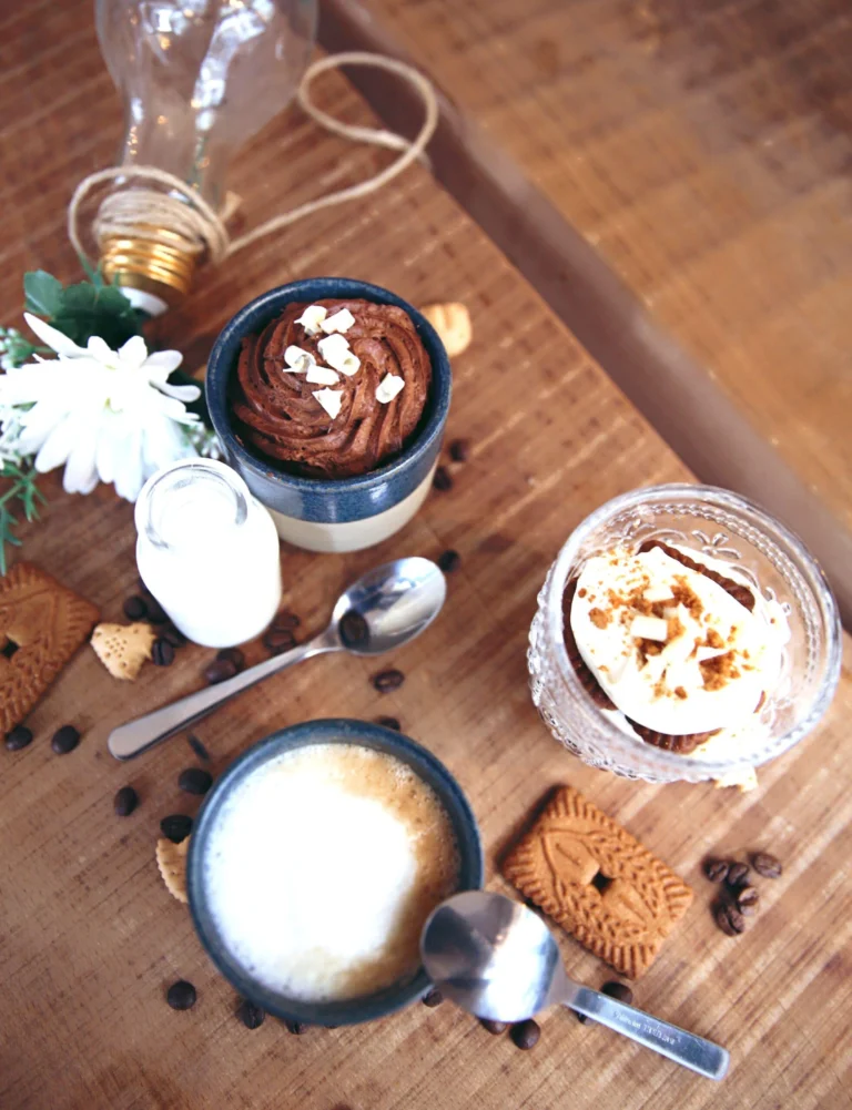 A wooden table seen from above with a milky coffee, a small pot of chocolate mousse, a glass dessert with crumble on top and speculoos biscuits