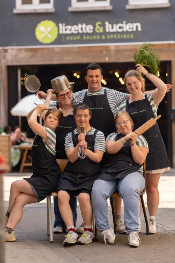 Six members of the Lizette & Lucien team in aprons, holding pans, a whisk and a rolling pin and posing under the lunch bar's shop sign