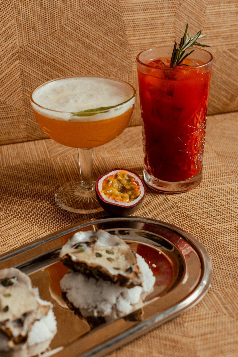 Two cocktails, one in a coupe with foam and one tall and red with a rosemary sprig, next to half a passion fruit and a silver tray of small bites