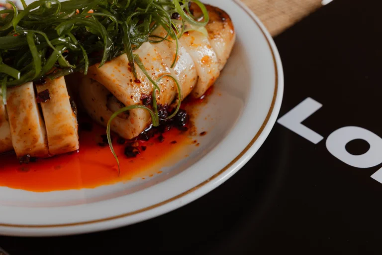 Close-up of a plated dish of sliced food glazed in red chilli oil and topped with spring onion, on a dark surface printed with the LOMA name