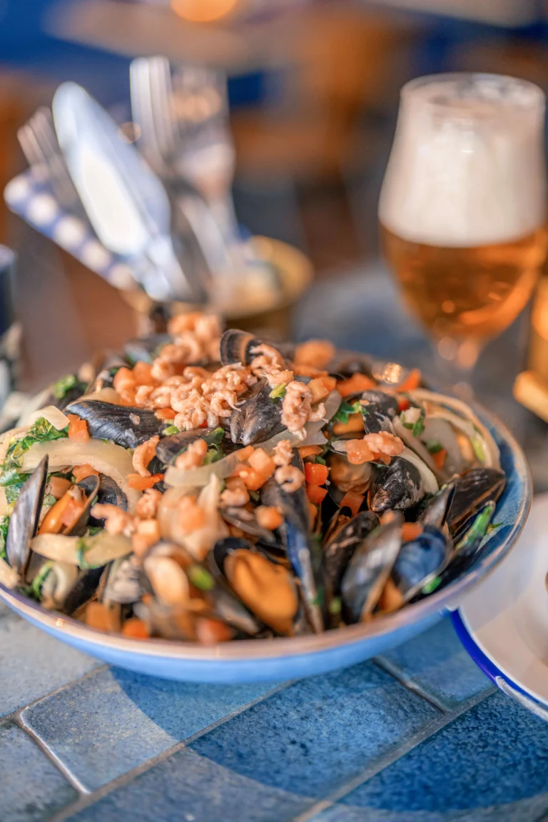 A blue-rimmed bowl of mussels in a creamy sauce topped with small shrimps, next to a glass of beer