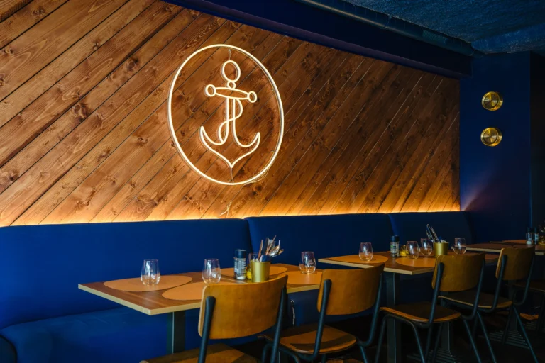 The dining room at Mac Moules, with a blue banquette, wooden tables and an illuminated anchor sign on a timber-clad wall