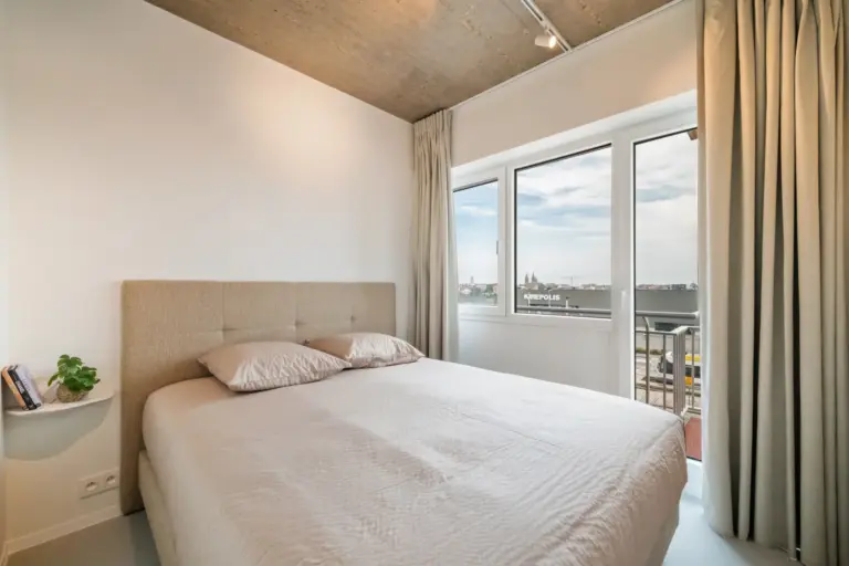 Calm double bedroom with an upholstered headboard, linen curtains and a corner window looking out over the town