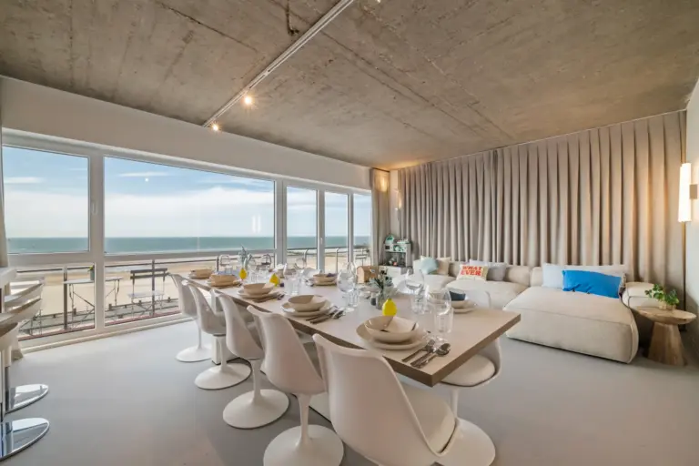 Open-plan living and dining room with a long laid table, white tulip chairs, a corner sofa and floor-to-ceiling sea-view windows