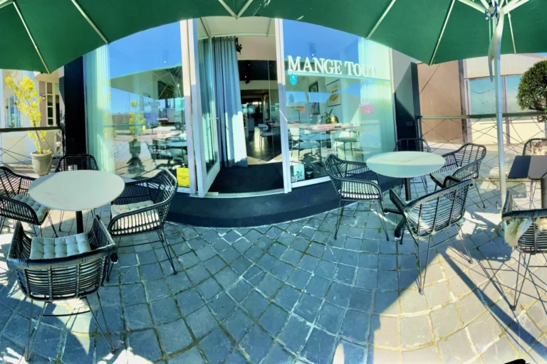 Terrace with green parasols, wicker chairs and round marble tables in front of the Mange Tout glass frontage