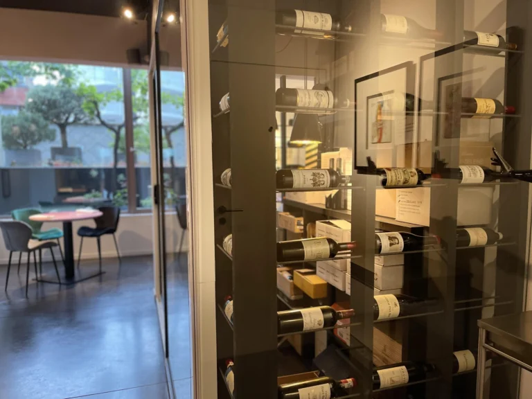 Glass-fronted wine wall with bottles and framed prints, a dining area with green chairs beyond