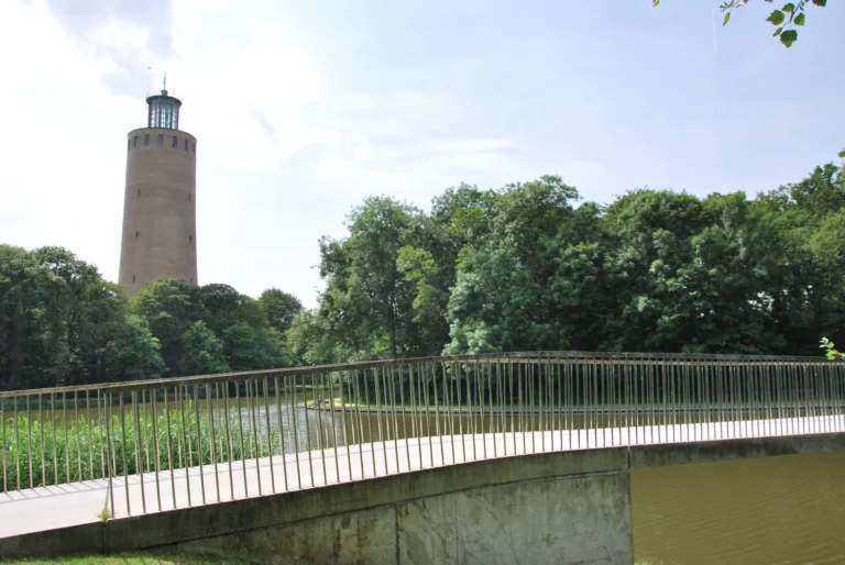 Slender footbridge with metal railings crossing the lake, the old water tower above the trees
