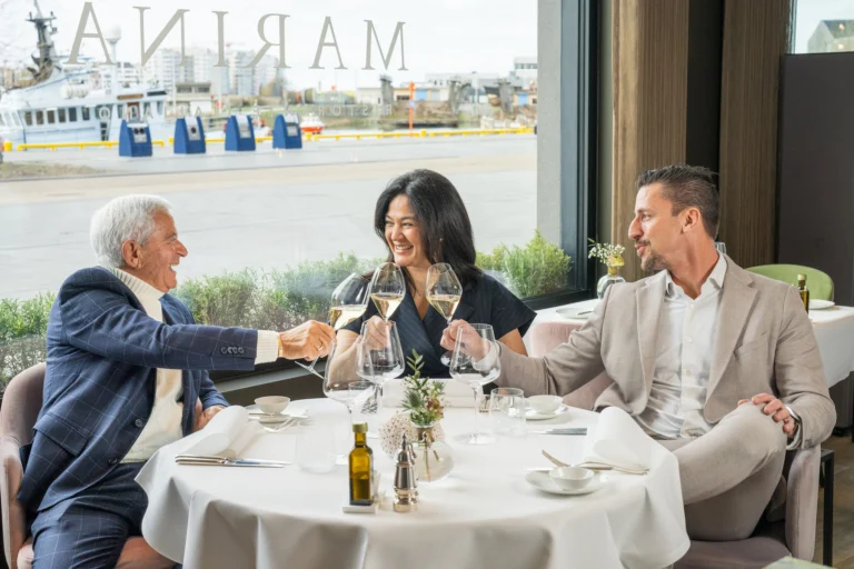 Three diners raising glasses at a window table, with the harbour and a fishing boat outside