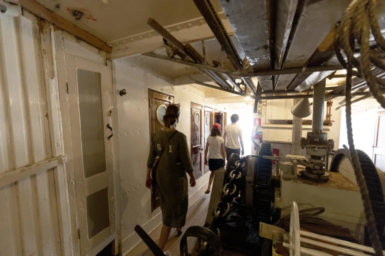 Visitors walking through a narrow below-deck passageway lined with white wooden cabins and ship machinery