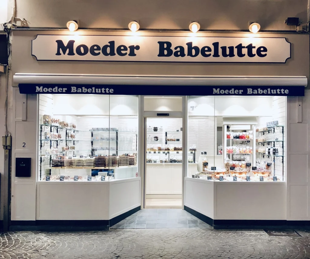 Moeder Babelutte shop front at dusk, with a lit sign above two large windows full of shelves of sweets and chocolate either side of an open glass door