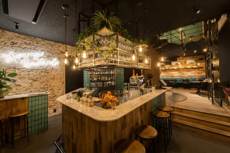 Marble-topped bar with wooden stools under a hanging glass rack with plants and Edison bulbs, next to a Mommy's Bastards neon sign on a stone wall