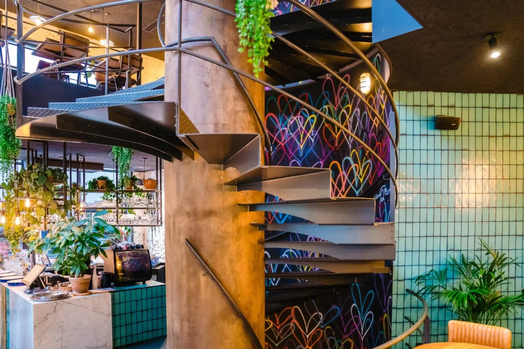 Steel spiral staircase in front of a wall painted with multicoloured hearts, with green tiles and plants around it