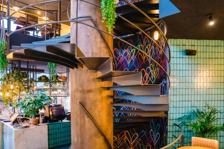 Steel spiral staircase in front of a wall painted with multicoloured hearts, with green tiles and plants around it