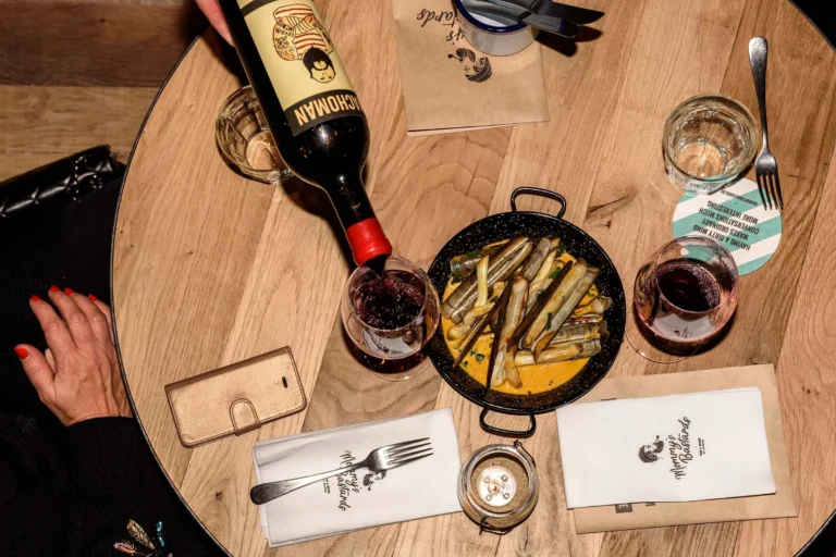 Overhead view of a wooden table with a pan of razor clams in sauce while red wine is poured