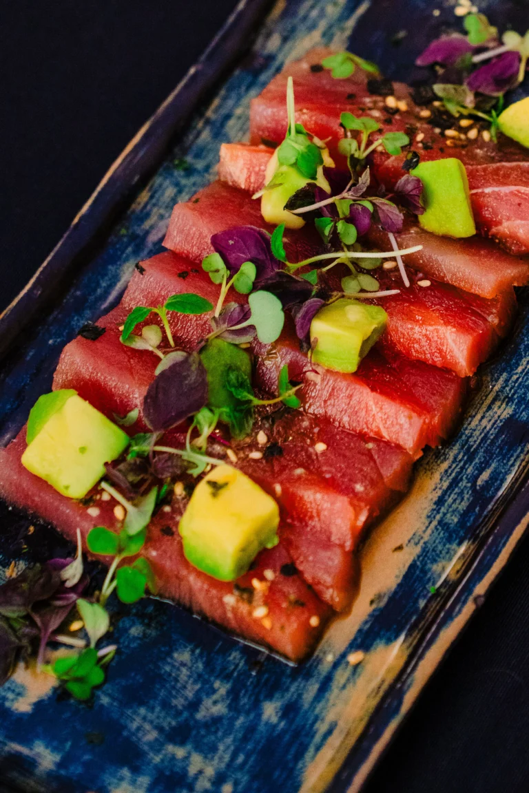 Slices of raw red fish topped with diced avocado and micro herbs on a blue patterned platter