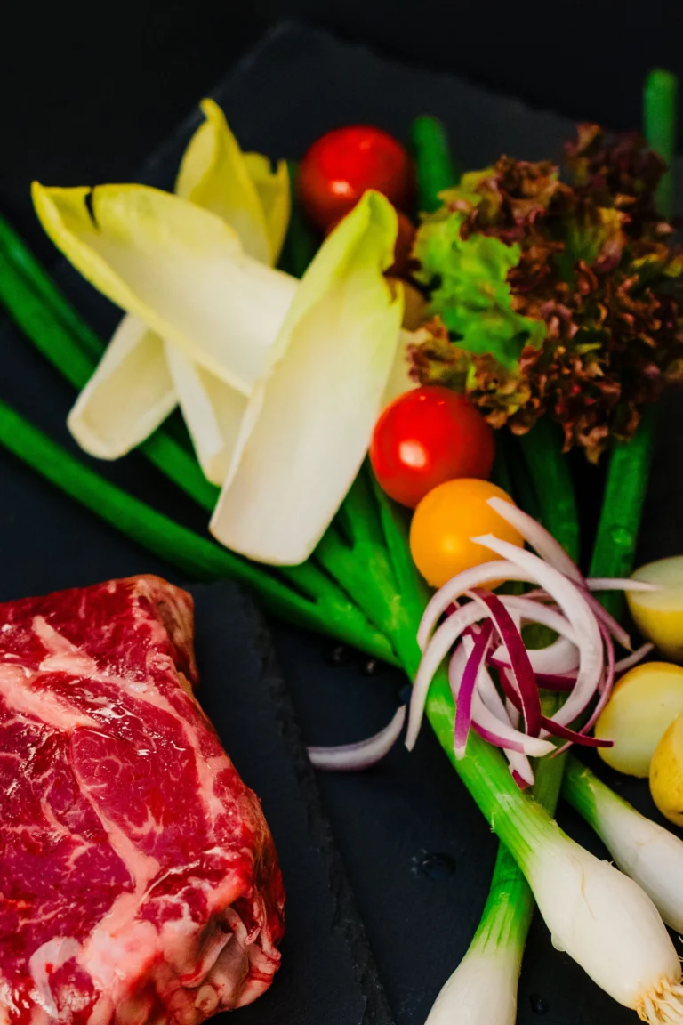 Raw marbled beef steak on dark slate beside chicory, spring onions, cherry tomatoes and red onion