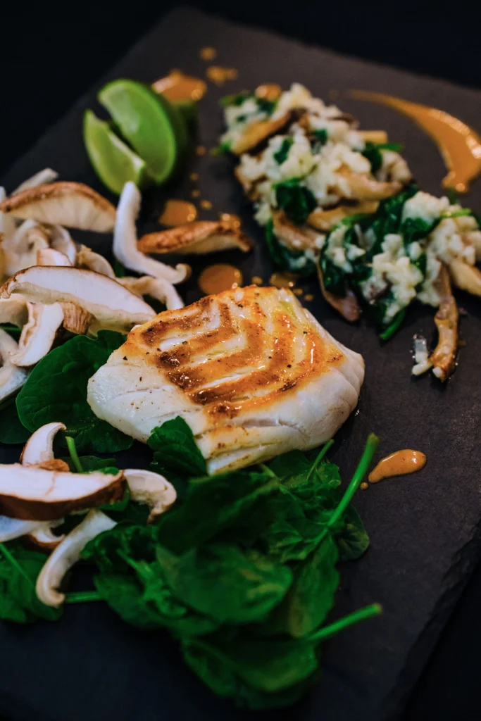 Grilled white fish fillet on a dark slate plate with sautéed mushrooms, spinach, a lime wedge and an orange sauce