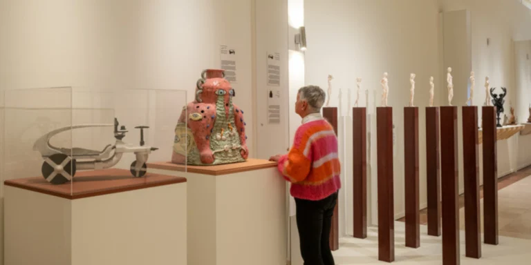 Visitor looking at a pink ceramic vase beside a row of small figurines on tall wooden plinths