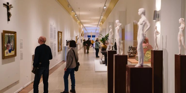 Long gallery lined with white statuettes on plinths and paintings, with visitors browsing the exhibition