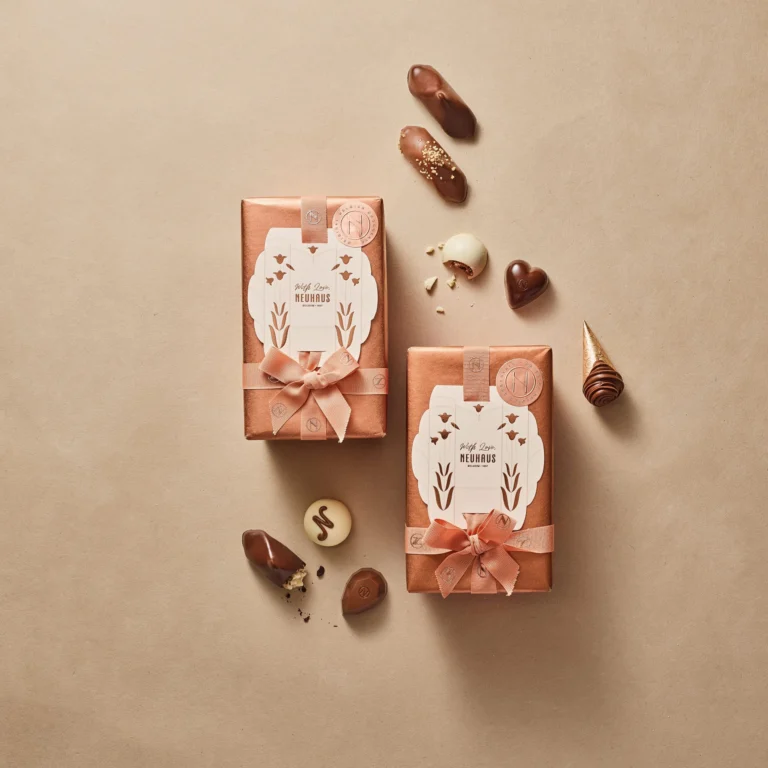 Two copper-coloured Neuhaus ballotins tied with ribbon bows, surrounded by scattered pralines on a beige background