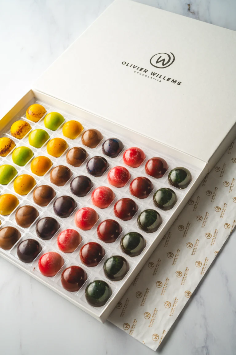 An open white Olivier Willems box holding rows of glossy pralines in yellow, red, green and brown on a marble surface
