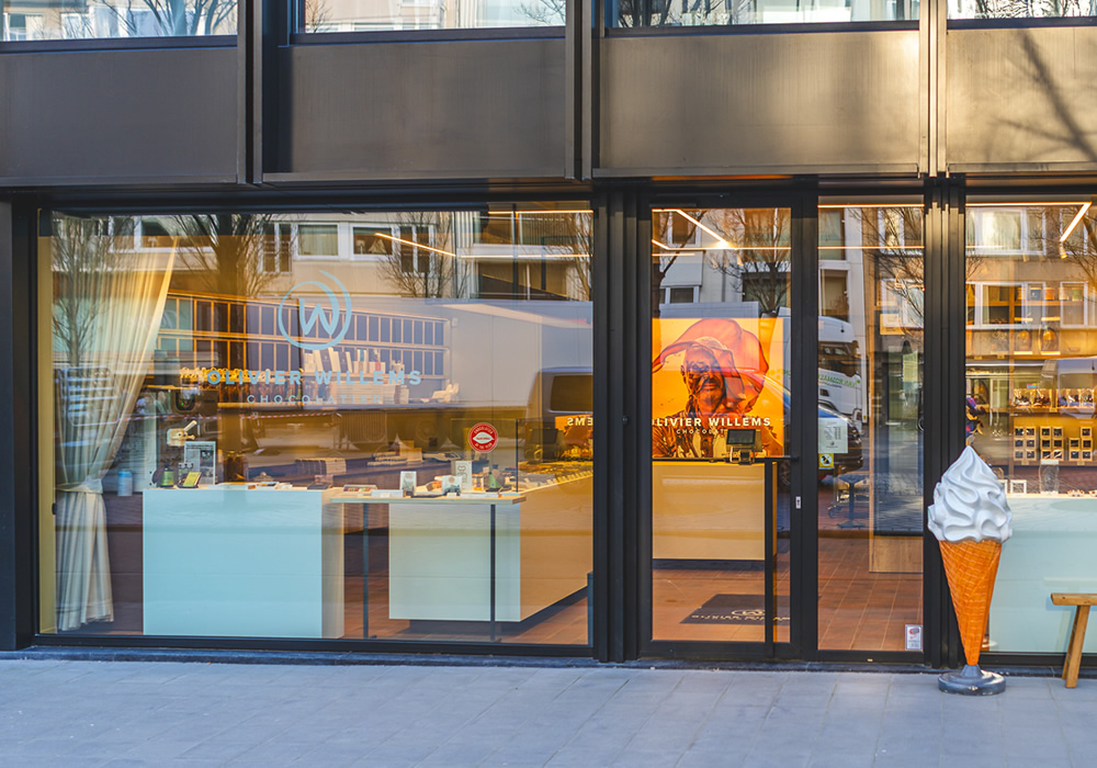 Glass shop front of Olivier Willems Chocolatier in a modern building, with the W logo in the window, a red poster on the door and a giant soft-serve cone outside