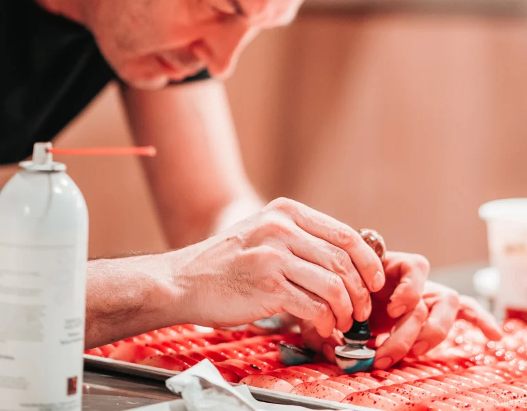 Close-up of a chocolatier's hands stamping a tray of red-speckled pralines in the workshop, with a spray can in the foreground