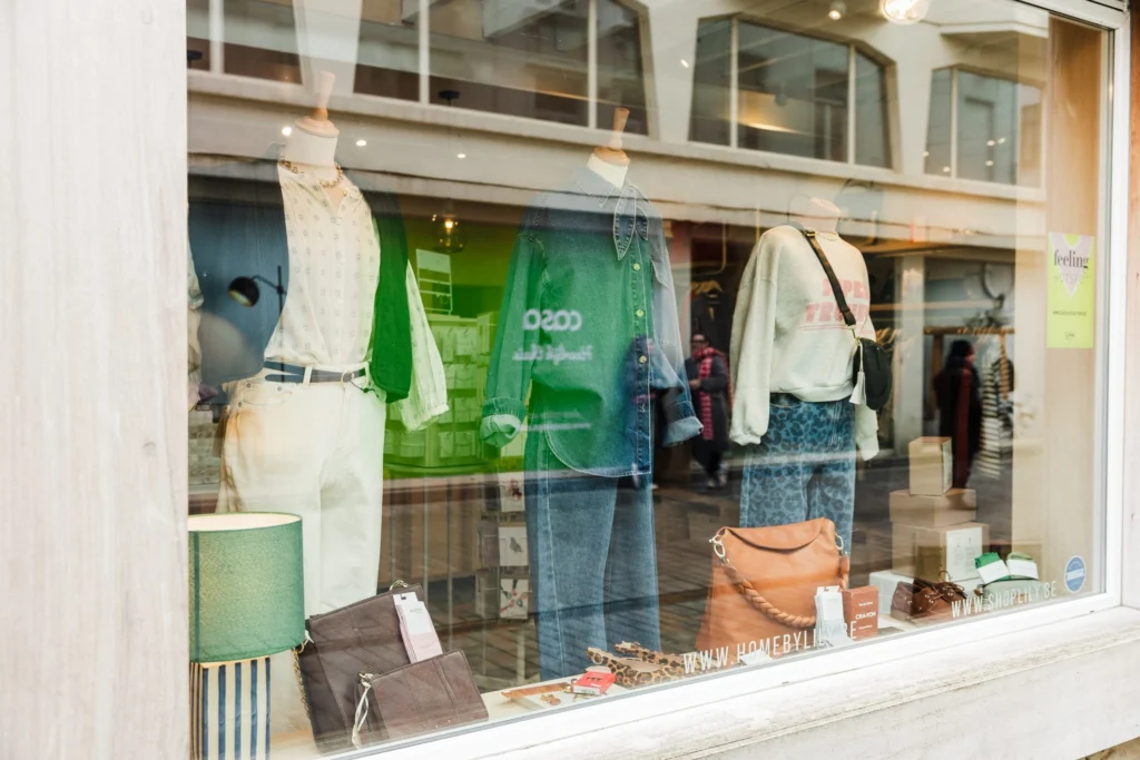 A boutique shop window in Ostend with mannequins in summer outfits, leather handbags on a shelf and a green table lamp, seen from the street