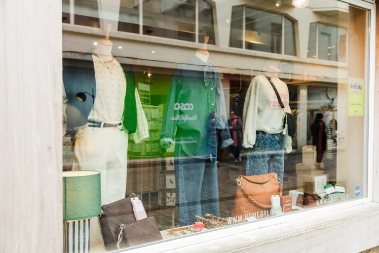 A boutique shop window in Ostend with mannequins in summer outfits, leather handbags on a shelf and a green table lamp, seen from the street