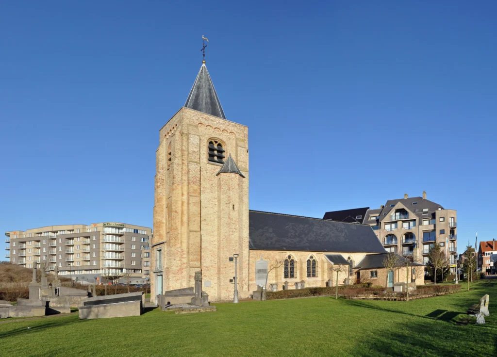 Small Gothic church with a square sandstone tower and a pointed slate spire, seen across the grassy churchyard under a blue sky