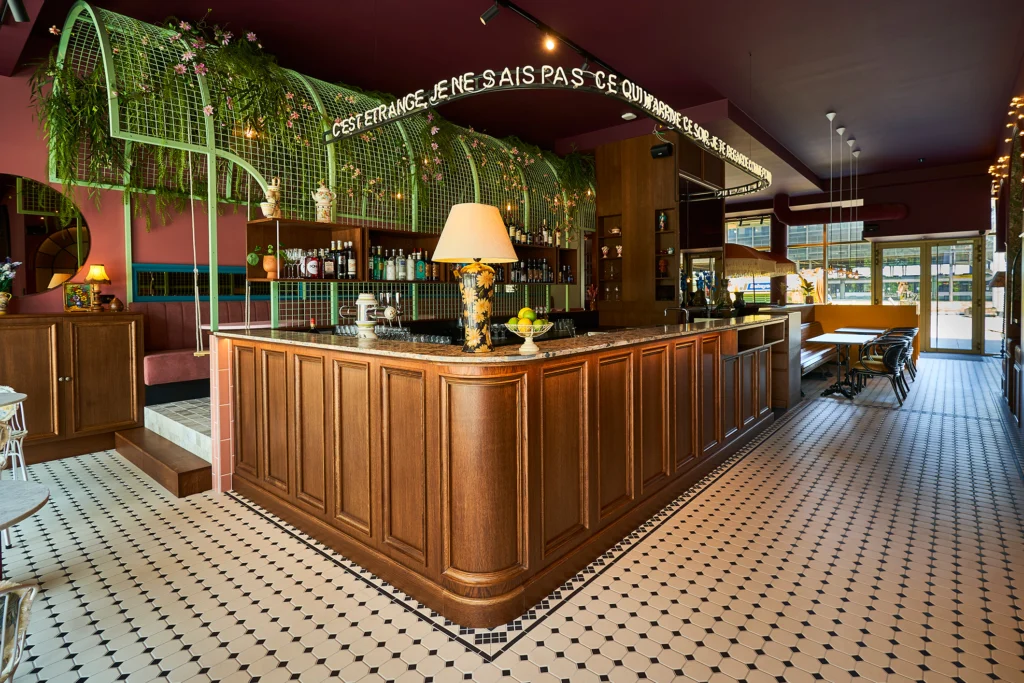 Long wooden bar under a green metal arch with hanging plants, a table lamp and a black-and-white mosaic floor