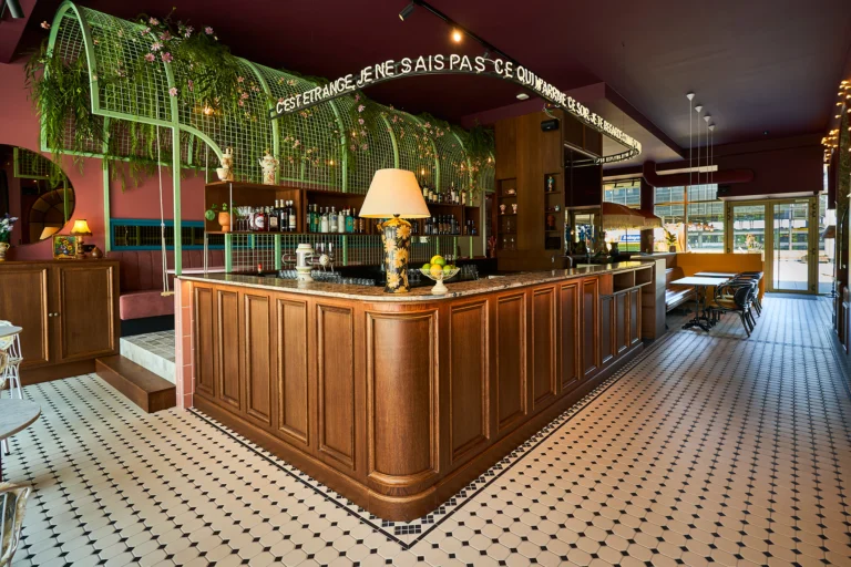 Long wooden bar under a green metal arch with hanging plants, a table lamp and a black-and-white mosaic floor