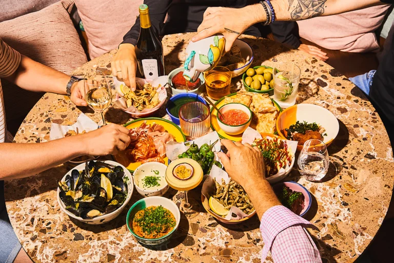 Overhead view of a terrazzo table covered with colourful small plates: mussels, charcuterie, padrón peppers, olives, fried fish and dips, with hands reaching in
