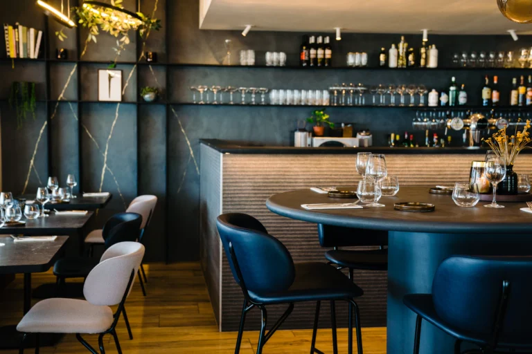 Dining room with a round dark table, blue and cream chairs and a bar with shelves of bottles and glasses behind