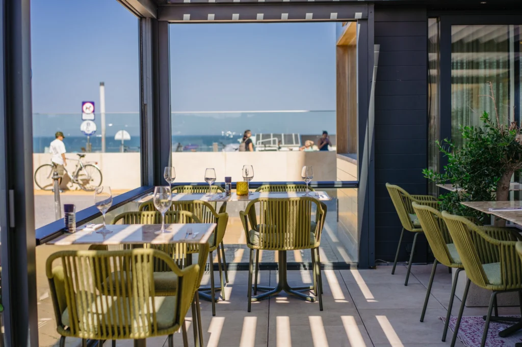 Terrace tables with olive-green woven chairs and wine glasses in the sun, with the beach, a cyclist and the sea visible through the open glass wall