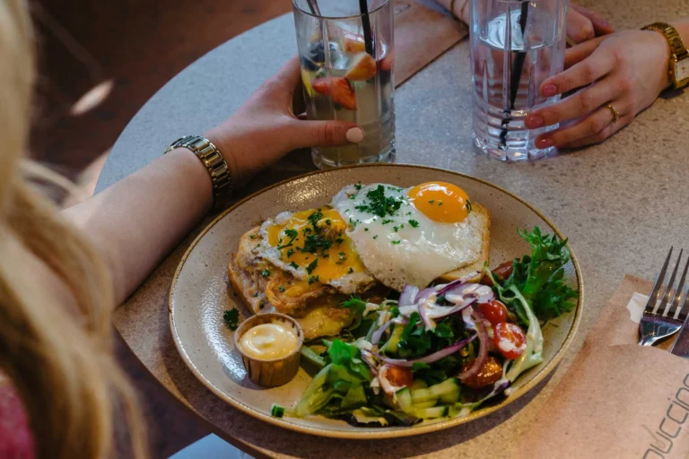 Brunch plate with fried eggs on toast, salad and a small pot of sauce, with glasses of water on the table