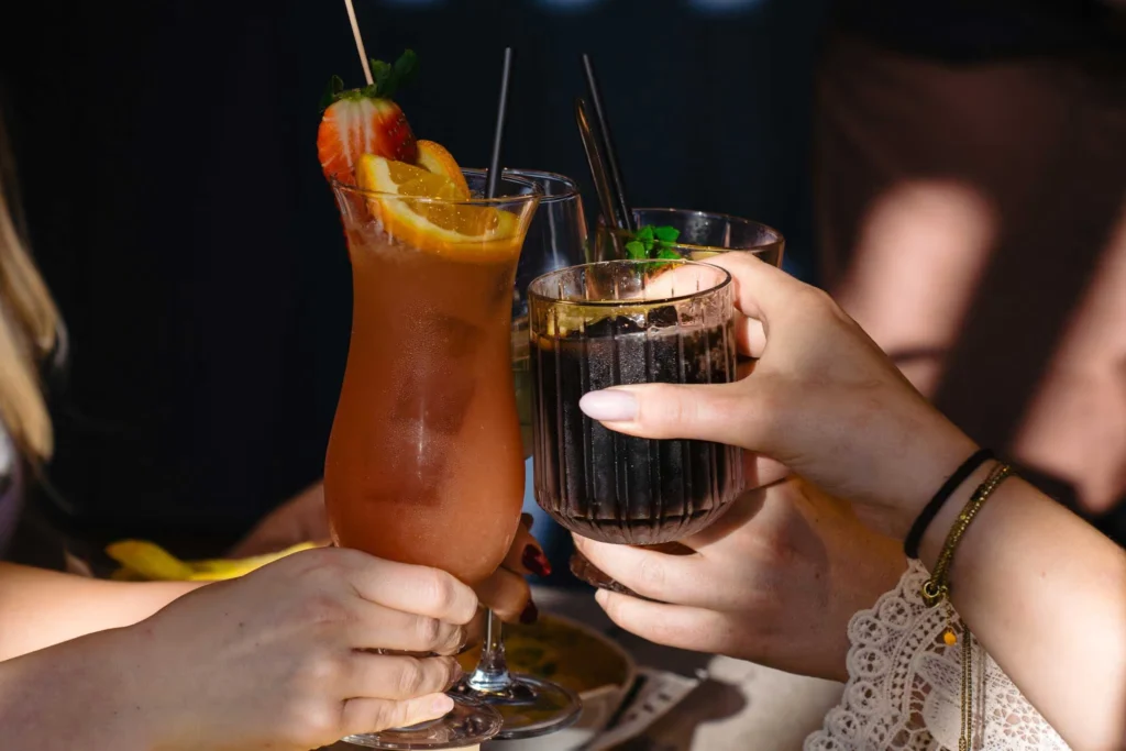 Hands raising a tall red cocktail garnished with strawberry and orange and a dark cocktail with mint in a toast