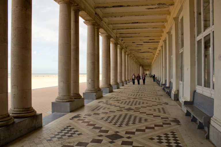 Covered walkway between columns with a patterned tiled floor, the beach visible on the left