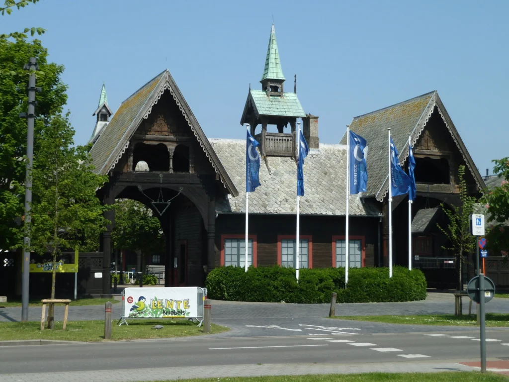 Dark timber gatehouse of the Royal Stables with two steep carved gables, a slate roof and a green copper turret, flags in front