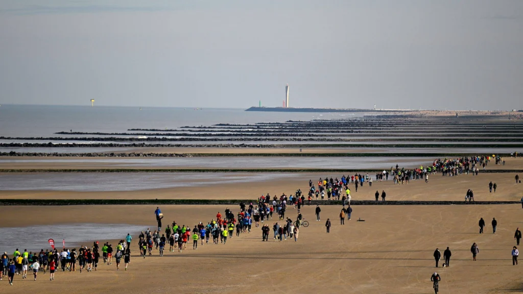 Long line of runners crossing a wide sandy beach at low tide, with breakwaters and a lighthouse on the horizon