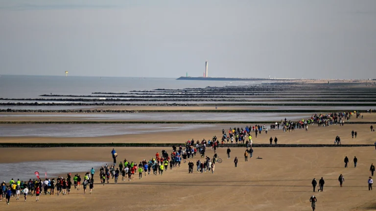 Long line of runners crossing a wide sandy beach at low tide, with breakwaters and a lighthouse on the horizon