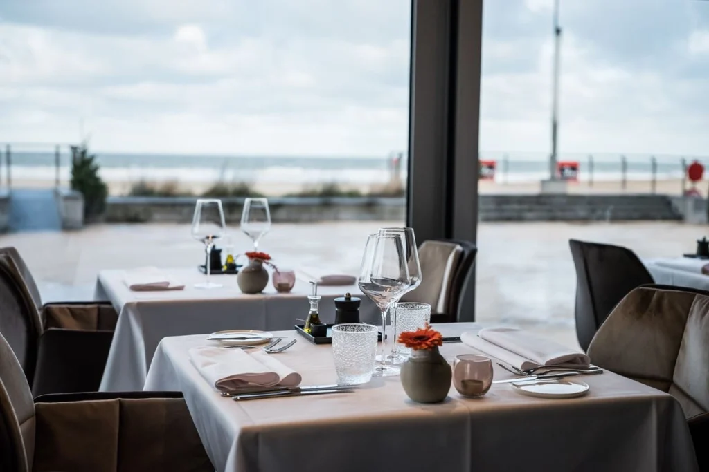 Laid tables with white cloths beside floor-to-ceiling windows overlooking the promenade and the sea