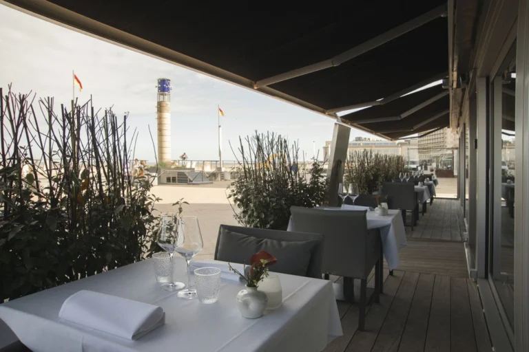Covered terrace with white-clothed tables on wooden decking, a beach tower and flags on the promenade beyond